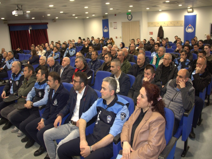 Bir konferans salonunda, mavi koltuklarda oturarak sahnede yapılan sunumu dinleyen zabıta personelleri ve sivil katılımcılar.