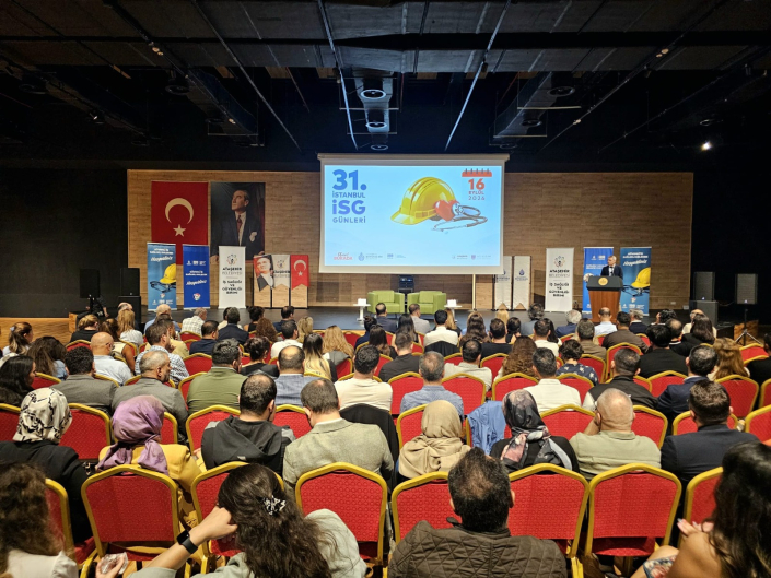 Fotoğrafta, bir konferans salonunda arkadan görünüşleriyle kırmızı koltuklarda oturan kalabalık bir dinleyici topluluğu ve karşılarındaki ışıklandırılmış sahnede kürsüde bir konuşmacı yer almaktadır. Projeksiyon perdesinde 31. İstanbul İSG Günleri, 16 Eylül 2026 yazmaktadır.