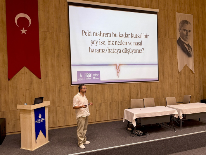 Bir konferans salonunda, sol tarafta Türk bayrağı, sağ tarafta Atatürk portresi asılıyken, bir erkek konuşmacı sahnenin merkezinde duruyor. Arkasındaki büyük ekranda, üzerinde İstanbul Büyükşehir Belediyesi (İBB) Sağlık logosu bulunan ve "Peki mahrem bu kadar kutsal bir şey ise, biz neden ve nasıl harama/hataya düşüyoruz?" yazılı bir sunum slaydının Türkçe metni gösterilmektedir.