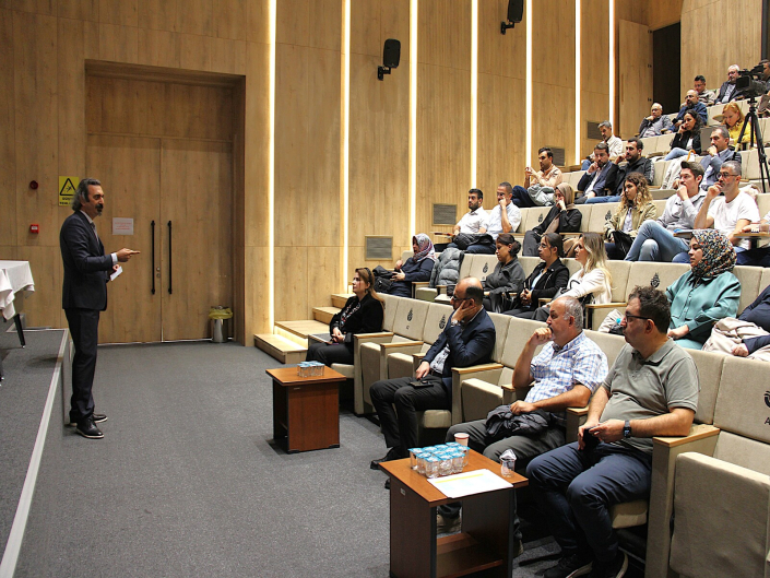 Modern ve ahşap panelli bir konferans salonunda, takım elbiseli bir erkek konuşmacı kürsüde sunum yaparken basamaklı sıralarda oturan dinleyiciler onu takip ediyor.