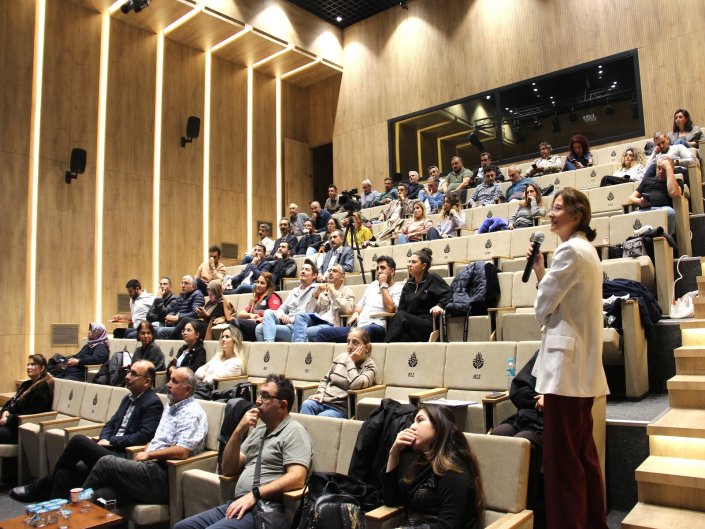 Modern ve ahşap panelli aynı konferans salonunda, beyaz ceketli bir kadın basamaklı sıraların önünde ayakta durarak elindeki mikrofonla konuşma yapıyor; salondaki diğer dinleyiciler ise projeksiyon perdesine bakarak sunumu takip ediyor.