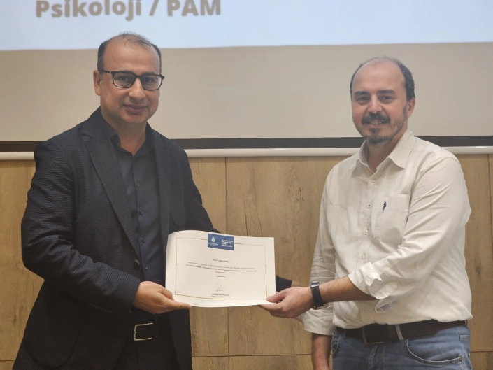 İBB İSG bünyesinde düzenlenen 'Yetişkin Psikolojisi ve Yetişkin Eğitiminde Etkin Yöntemler' konulu sempozyumda sertifika takdim anı. Arka planda 'Psikoloji / PAM' yazılı ekranın önünde, takım elbiseli bir yetkili, beyaz gömlekli konuşmacıya İstanbul Büyükşehir Belediyesi logolu teşekkür belgesini sunuyor.