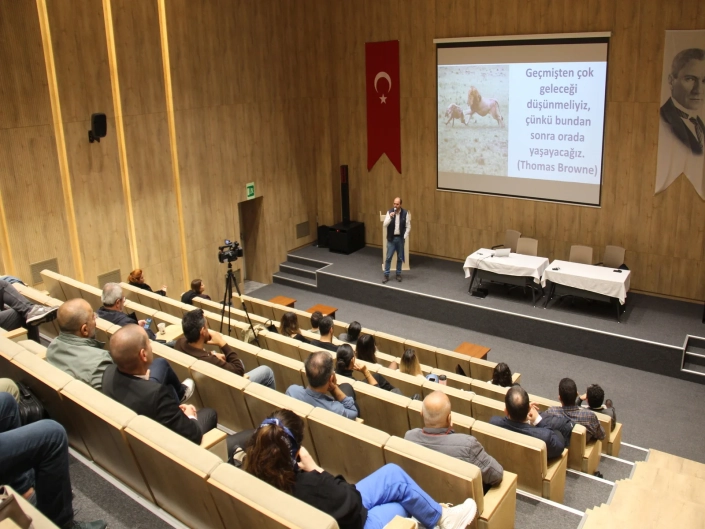 İBB İSG'nin 'Yetişkin Psikolojisi ve Yetişkin Eğitiminde Etkin Yöntemler' sempozyumunun gerçekleştirildiği geniş, ahşap kaplamalı amfi salonu. Sahnedeki kürsüde mikrofonla konuşan bir eğitmen ve arkasındaki büyük projeksiyon ekranında Thomas Browne'un gelecek temalı sözü yer alıyor; basamaklı koltuklarda katılımcılar sunumu dinliyor.