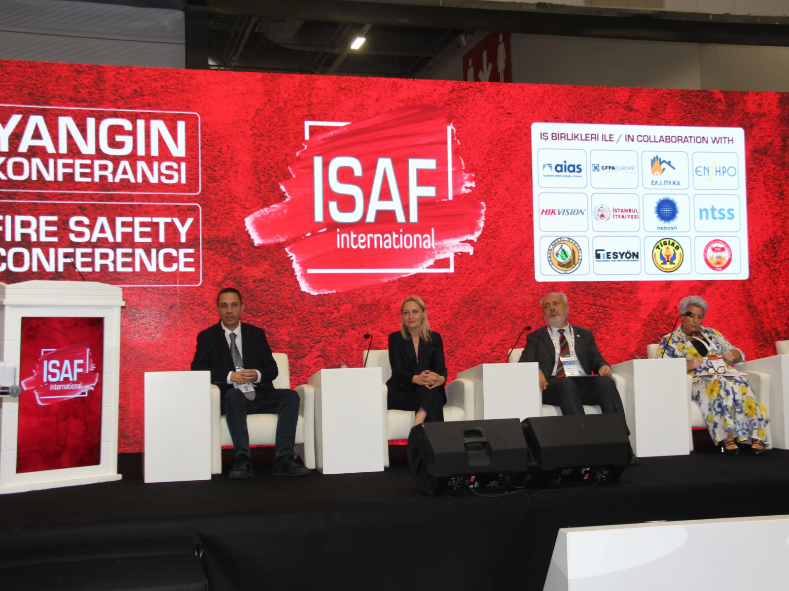 ISAF International Fire Safety Conference (Yangın Konferansı) sahnesinde, arkalarında iş birliği yapılan kurumların logolarının bulunduğu kırmızı bir fon önünde, yan yana dizilmiş beyaz koltuklarda oturan dört konuşmacı (takım elbiseli iki erkek, siyah ceketli bir kadın ve desenli elbiseli bir kadın) yer alıyor.