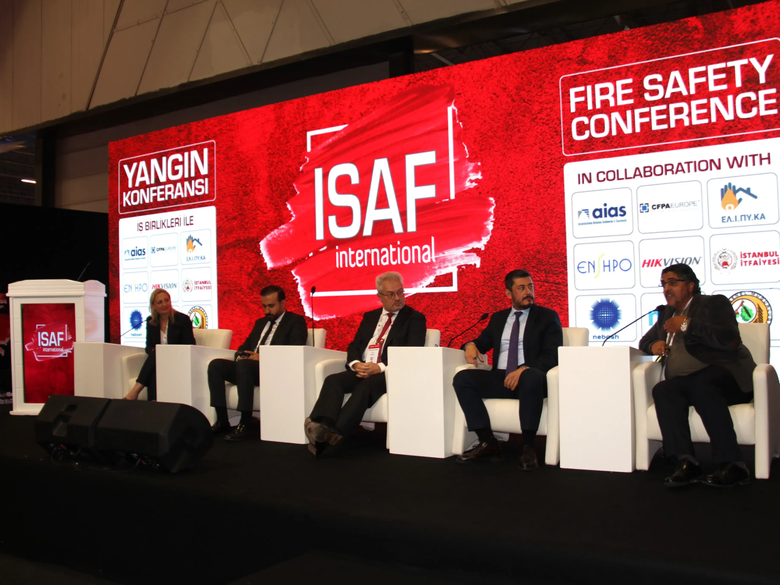ISAF Fire Safety Conference (Yangın Konferansı) sahnesinde, üzerinde ISAF logosu ve iş birliği ortaklarının amblemleri bulunan büyük kırmızı bir ekranın önündeki beyaz koltuklarda yan yana oturan biri kadın beş panelist konuşma gerçekleştiriyor.