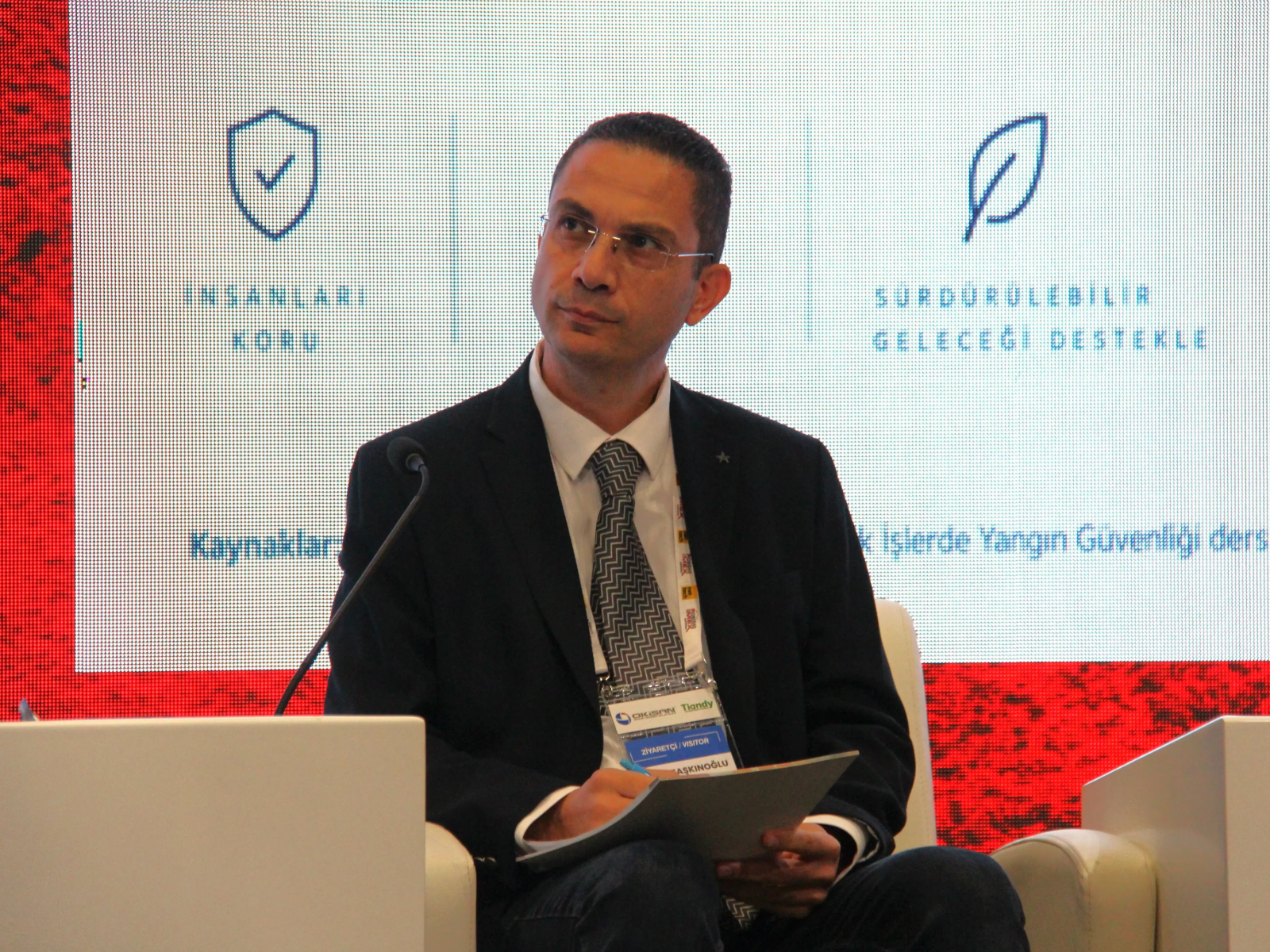 Bir konferansta elinde kağıtlar tutarak oturan gözlüklü ve takım elbiseli bir erkek konuşmacı arkasındaki dijital ekranda "İnsanları Koru" ve "Sürdürülebilir Geleceği Destekle" yazılı simgeler önünde dinliyor.